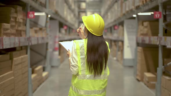Back view. Female Worker Wearing Hard Hat Checks Stock and Inventory Walks in the Retail Warehouse 