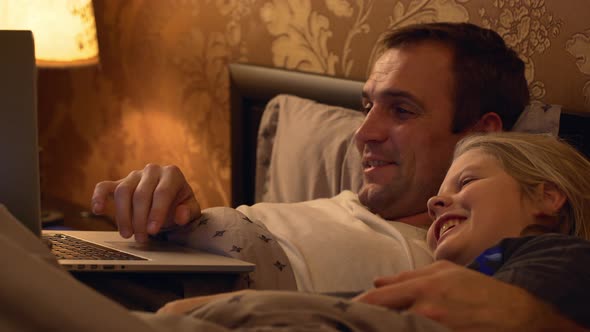 Footage of Middleaged Father with Light Bristle Lying in Bed with Laptop and His Small Blonde Child