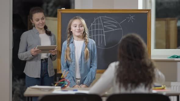 Portrait of Smart Cute Caucasian Schoolgirl Talking and Smiling Standing in Classroom with Teacher