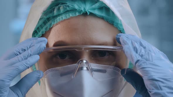 Young Woman Doctor in Hazmat Suit Puts on Medical Glasses and Protective Mask