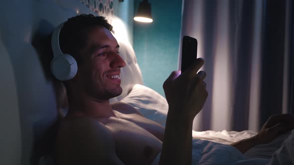 Smiling Man in Headphones Listening to Music and Using Smartphone While Lying in Bed in Bedroom at