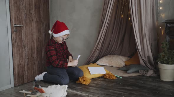 Caucasian Boy Wearing Glasses and a Santa Cap Puts a Letter in a Craft Envelope