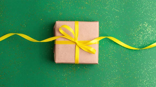Brown gift box with a yellow satin ribbon bow on jade green background with gold colored glitter