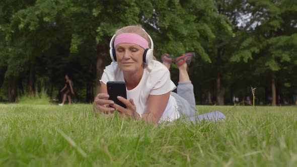 Senior Woman in Sportswear with Earphones Listening To Music on Smartphone Lying on Grass at Park