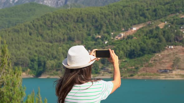 Woman in Hat with Wild Hair Shooting a Video on Mobile Phone of Mountains Lake Background