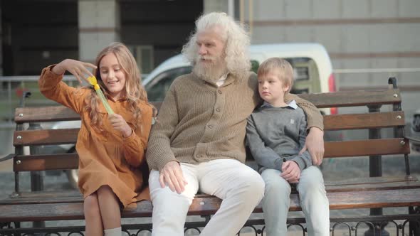 Relaxed Caucasian Grandfather and Grandson Sitting on Bench As Granddaughter Blowing Soap Bubbles