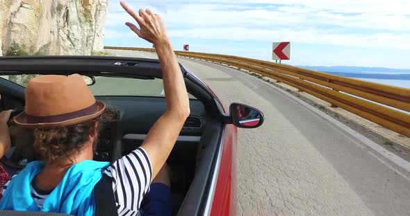 Hipster man with hat waving arms driving on winding coastal road in convertible