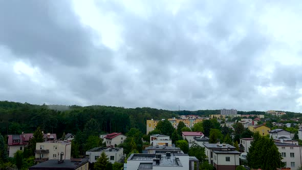 Time lapse shot of grey clouds flying over rural town with colorful houses beside forest trees