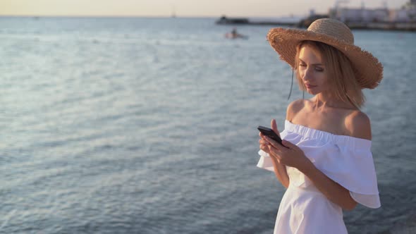 Young Caucasian Woman Walks By the Sea or Ocean Early in the Morning at Dawn Holding a Smartphone in