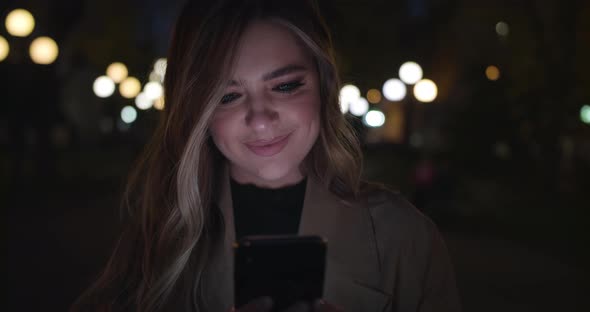 Cute Girl Uses Smartphone and Smiles Standing on Night Street in the City. Woman Messaging on Phone