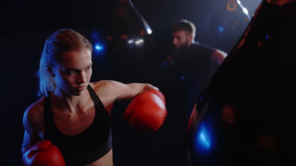 Strong Woman is Punching the Boxing Bag Wearing Red Gloves and the Man is Training on the Background