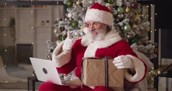 Cheerful Santa Claus Sitting on an Armchair Against a Christmas Tree Background in His Residence