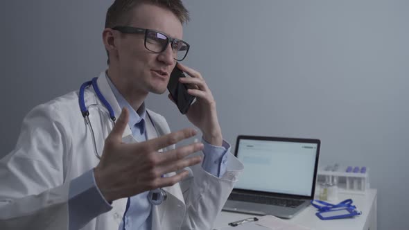 European Doctor in White Medical Coat and Glasses Consults Patient on Cell Phone in Clinic Office