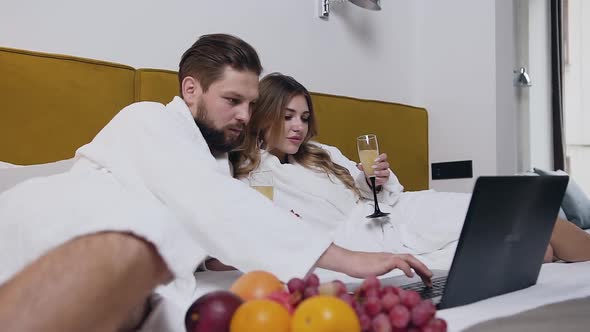 Young Couple in white Bathrobes Lying on Big Bed in Hotel and Watching Together at Computer