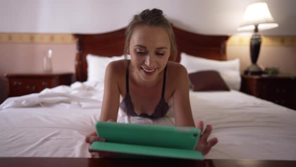 Front View Portrait Happy Relaxed Young Woman in Bra Lying on Bed Surfing Internet on Tablet Smiling