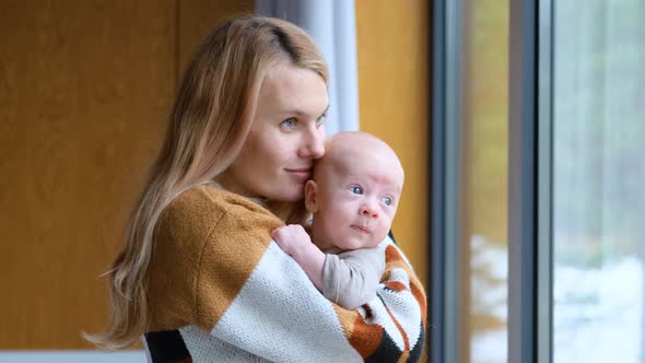 woman hugs and kisses toddler boy standing by the window