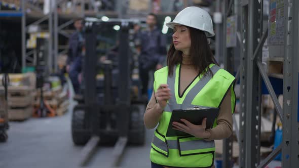 Concentrated Female Freight Agent in Hard Hat Standing in Warehouse Filling in Bill of Lading As Men