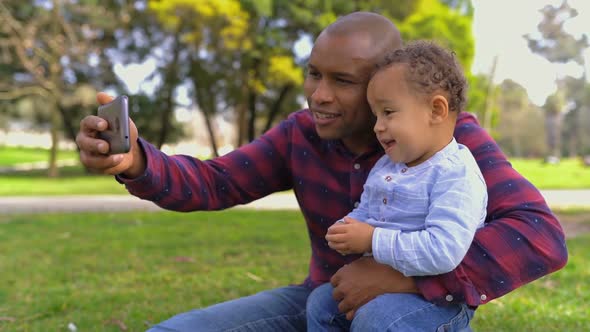 Father Sitting on Haunches, Holding Son on Knees, Making Selfie