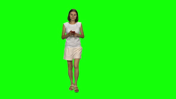 Young Woman Is Calmly Walking and Texting Message Vie Her Mobile Phone on Green Screen. Front View.