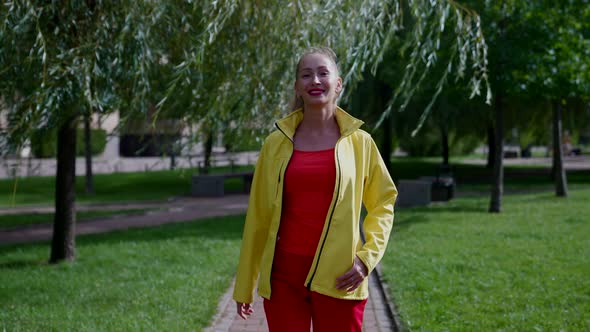 a Woman in Red Clothes and a Yellow Jacket Walks Along a Path in a City Park