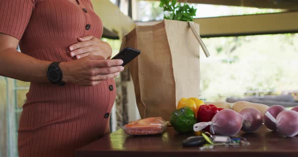 Midsection of happy caucasian pregnant woman touching belly, using smartphone in kitchen