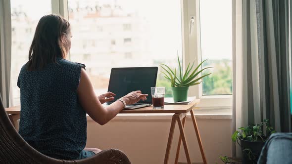 A Woman Working on a Laptop While Sitting at the Table Opposite a Window