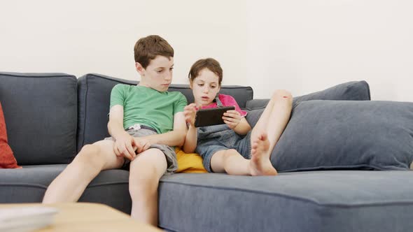 Two kids sitting on a large sofa at home, using a mobile phone