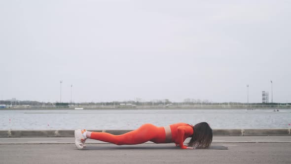 A beautiful woman in a bright orange fitness suit makes exercises on the street. A young girl plays