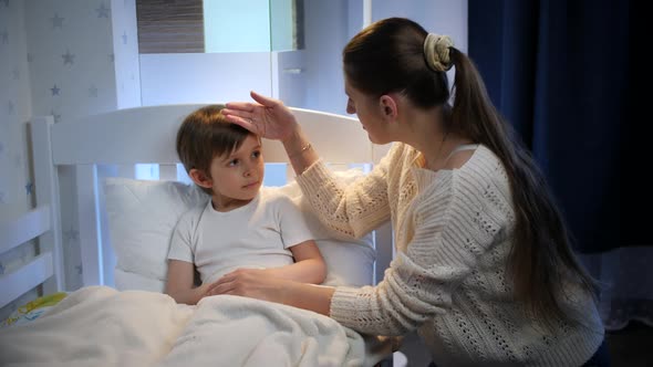 Young Caring Mother Touching Forehead of Her Sick Little Son Lying in Bed