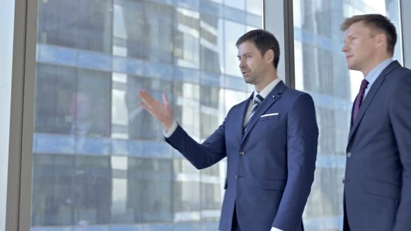 Two Middle Aged Businessmen Having Discussion and Pointing Outside the Window