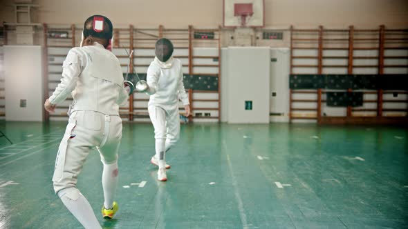 Two Young Women in Protective Costumes and Helmets at the Fencing Training in the School Gym