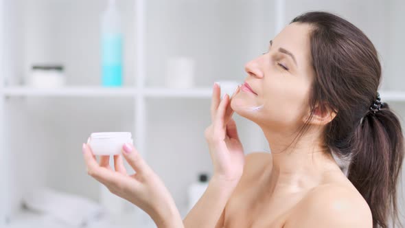 Young Woman Smiling and Enjoying Applying Antiageing Moisturizing Cream on Perfect Skin Face