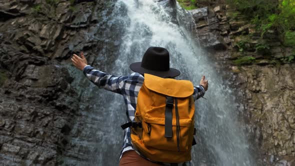 Girl Traveler Wearing a Hat and a Yellow Backpack While Standing Against a Waterfall