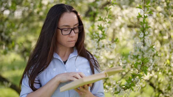Adult Woman with Glasses for Vision is Reading Book Standing in Blooming Garden in Spring Day