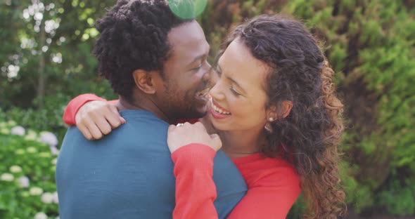 Happy biracial man hugging his fiance with engagement ring on hand in garden in sun