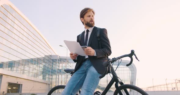 Businessman in Formal Clothes which Working with Financial Reports, Leaning on His Bicycle