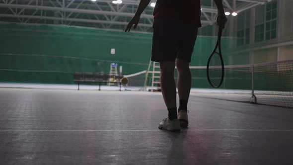 Male Tennis Player is Walking on Indoor Court Throwing and Catching Ball Holding Racquet Rear View