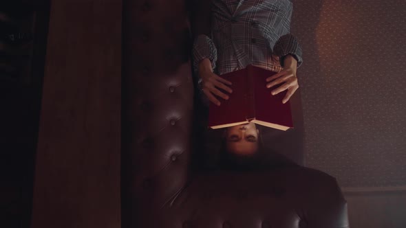 Young Attractive Caucasian Woman Holds a Book in Her Hands and Reads It While Lying on the Couch in