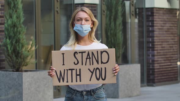 Caucasian Female Protester Activist Wearing a Medical Mask Holds a Cardboard Poster with the Slogan
