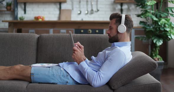 Handsome Businessman in Headphones Lying in Cozy Couch Looking Document Paper in Modern Kitchen