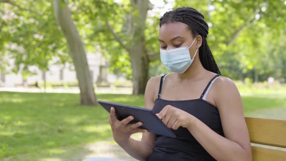 A Young Black Woman in a Face Mask Works on a Tablet and Sits on a Bench in a Park on a Sunny Day