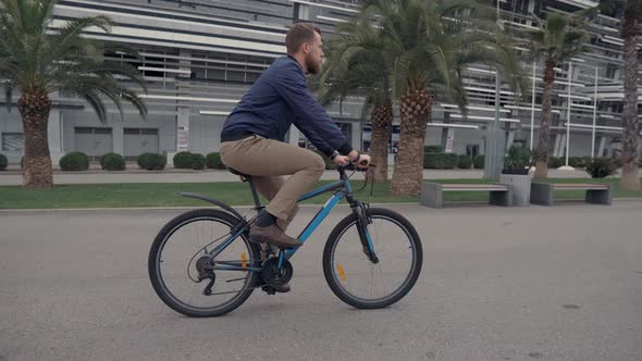 Young Bearded Man Is Riding Bicycle in Park Area in Summer Day Side View