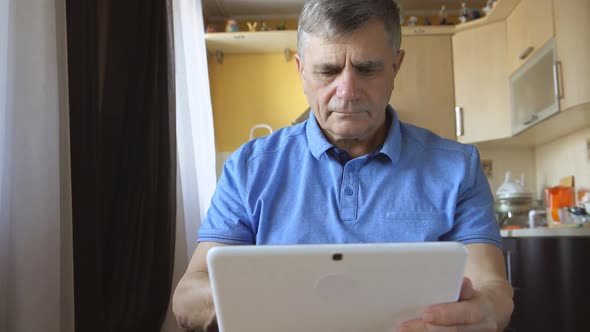 Portrait Of A Senior Man At The Blue Shirt Sits And Types A Tablet Pc At Home