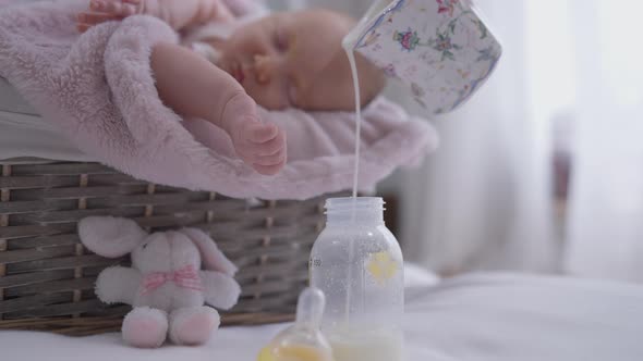 Pouring Milk in Baby Bottle with Blurred Infant Sleeping at Background in Baby Basket