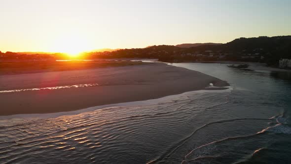 Aerial flight over nature reserve with calm water and beach at Mangawhai Heads with golden sunset in