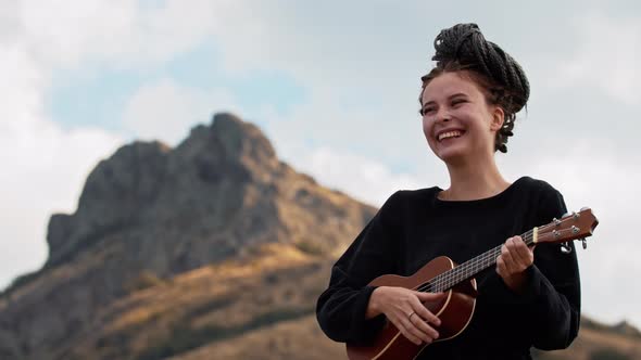 Young Laughing Woman with Dreadlocks Tied Up to Her Head Playing Ukulele in Mountains