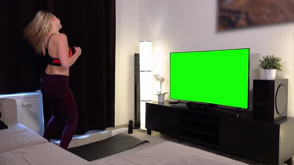 A young beautiful woman does a fitness dance in front of a TV with a green screen in an apartment