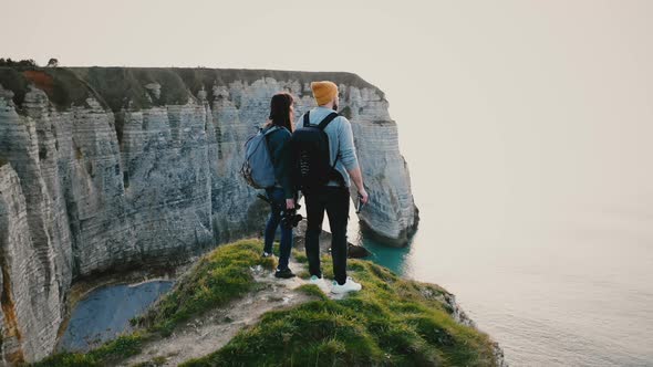 Drone Flying Around Young Happy Tourist Couple with Backpacks Watching Epic Sunset Sea on Top