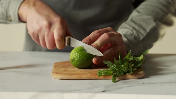 Bartender is Cutting a Juicy Green Lime on a Chopping Board with a Knife and a Sprig of Mint is