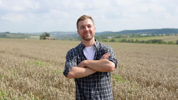 Happy Young Farmer Standing on Wheat Field While Combine Harvester in Background